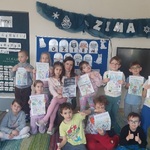 Dzieci, razem z Panią Wioletą siedzą pod tablicą u trzymają rysunki. 
