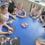 Dzieci siedzą przy stolikach i wykonują prace kolorową kredą na czarnym papierze.