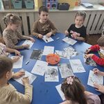 Dzieci siedzą przy stoliku i rysują węglem kreślarskim na białych kartkach.