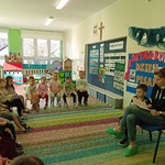 Przedszkolaki siedzą na krzesełkach i słuchają pani_ która czyta ksiażkę.