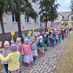 Dwie grupy dzieci stoją parami przed budynkiem przedszkola_ unoszą papierowe kwiaty.