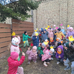 Dzieci z dwóch  grup stojąc lub kucając na tle muru podnoszą w górę papierowe kwiatki.