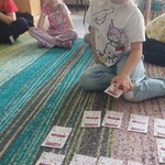 Dziewczynka siedzi na dywanie_ przed nią leży memory_ trzyma w dłoni jedną kartę_