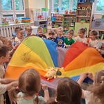 Dzieci stoją w kole_ w dłoniach trzymają kolorową chustę_ Podrzucają misiem, który jest na chuście_