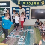 Na dywanie rozłożona gra. Chłopcy stoją na jednej nodze, dziewczynki siedzą i się przyglądają. 