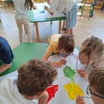 dwie grupki dzieci przy stolikach rozdmuchują farbę za pomocą słomek.