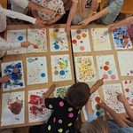 Grupa przedszkolaków stoi wokół stolików z pracami plastycznymi wskazując własne dzieła.