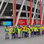grupa dzieci w kamizelkach bezpieczeństwa trzyma transparent z nazwą przedszkola na tle stadionu.