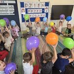 Dzieci siedzą na dywanie w kole i trzymają w górze balony.