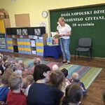 Pani w białej bluzce stoi naprzeciwko dzieci i trzyma otwartą książkę.