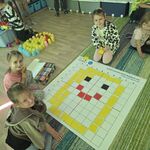 Trzy dziewczynki siedzą na dywanie i prezentują uśmiech ułożony z kolorowych kartoników na macie do kodowania.