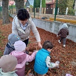 Kilkoro dzieci wrzuca liście do worka trzymanego przez opiekunkę.