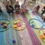 Dzieci siedzą na dywanie a przed nimi leżą przedmioty w kolorowych obręczach.