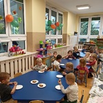Grupa przedszkolaków siedzi przy stolikach i je ciasto.