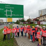 grupa dzici przedszkolnych w pomarańczowych kamizelkach odblaskowych, pod znakiem drogowym  informującym o skrzyżowaniu dróg i kierunkach