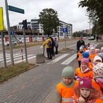 grupa dzieci w pomarańczowych kamizelkach , w pobliżu przejścia dla pieszych. W tle ulica