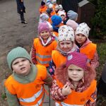 grupa dzieci w pomarańczowych kamizelkach idąca na spacer. Na pierwszym tle chłopiec zaglądający w obiektyw