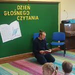 na zdjęciu klęczący mężczyzna czytajacy dzieciom ksiązkę. z tyłu żólty napis dzień głosnego czytania.jpg