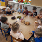 grupa dzieci siedzi wokół stołów na których znajdują się gruszki, śliwki, szarlotka. Dzieci pijąsok