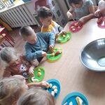 Dzieci siedzące z lewej strony stolika, kroją owoce na kolorowych talerzykach