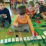 Dzieci siedzą na dywanie. Chłopiec przy pomocy kart do kodowania zaprogramowuje robota Genibot 