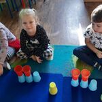Dwie dziewczynki i chłopiec prezentują wykonanie zadania z kolorowych kubeczków