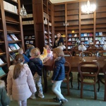Dzieci chodzą w pomieszczeniu czytelni, wszędzie wysokie półki na czasopisma, po prawej stronie duży stół z lampą.jpg