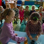 Grupa dzieci obserwuje jak dwie dziewczynki wsypują do wody barwnik spożywczy
