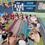 Grupa dzieci siedzi w kole na dywanie z misiami, trzymają w ręku drewnianą łyżkę.jpg