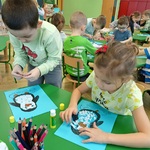 Grupa dzieci siedzi przy stolikach i wykleja papierkami sylwetę pingwina