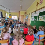 Dzieci siedzą na krzeslach i dywanie patrząc na tablice.