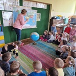 Pani stoi przy mapie świata _ obok na dywanie znajduje się globus.