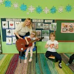 siedząca na krześle dziewczynka próbuje grać na gitarze z pomocą kobiety 