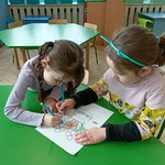 Dwie dziewczynki siedzą przy stoliku i kolorują rysunek przedstawiający kwiaty w wazonie
