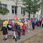Grupa dzieci z własnoręcznie wykonanymi kwiatami i gaikiem stoi przed budynkiem przedszkolnym