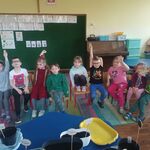 sala przedszkolna _ dzieci siedzące na krzesłach z podniesioną ręką.