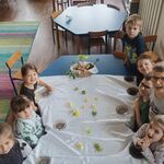 dzieci siedzą przy stole przykrytym białym obrusem_ na środku rozsypane pisanki_ u góry ozdobiony koszyczek - święconka.