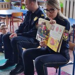 strażnicy miejscy siedzą na krzesłach. Strażniczka pokazuje dzieciom książkę.