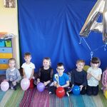 grupa dzieci siedzących na kolorowym dywanie_ każde trzyma kolorowy balon. balony tworzą napis FUNDACJA A JAK.