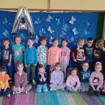 grupa uśmiechniętych dzieci ustawiona przy niebieskiej tablicy, na której przyczepione są niebieskie motyle i balon w kształcie litery A