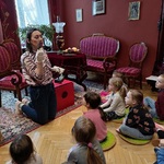 Dzieci siedzą na poduszkach i oglądają eksponat prezentowany przez panią przewodnik.