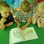 Troje dzieci siedzi przy stoliku i ogląda książkę.