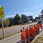 Dzieci ustawione parami stoją na chodniku przy ruchliwej ulicy; z lewej strony widoczny znak drogowy.