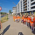 Dzieci ustawione parami stoją na szerokim chodniku obok znaku drogowego.