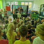 Dwaj aktorzy trzymają w ręku pacynki_ obok stoi sylweta wozu policyjnego.