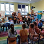 Dzieci siedzą na krzesełkach i słuchają czytanej książki.