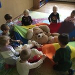 8 dzieci siedzi wokół chusty animacyjnej trzymając ją za uchwyty_ w środku chusty leżą misie.