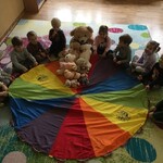 10 dzieci siedzi w półkolu wokół chusty trzymając ją za uchwyty_ na małym skrawku chusty ułożone są pluszowe misie.