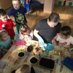 dzieci zgromadzone wokół stołu oglądają ziarenka sypane na talerzyk przez kobietę.