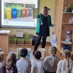 Kobieta stoi przed dziećmi_ w jednej ręce trzyma torbę_ a w drugiej żółty pojemnik na odpady.